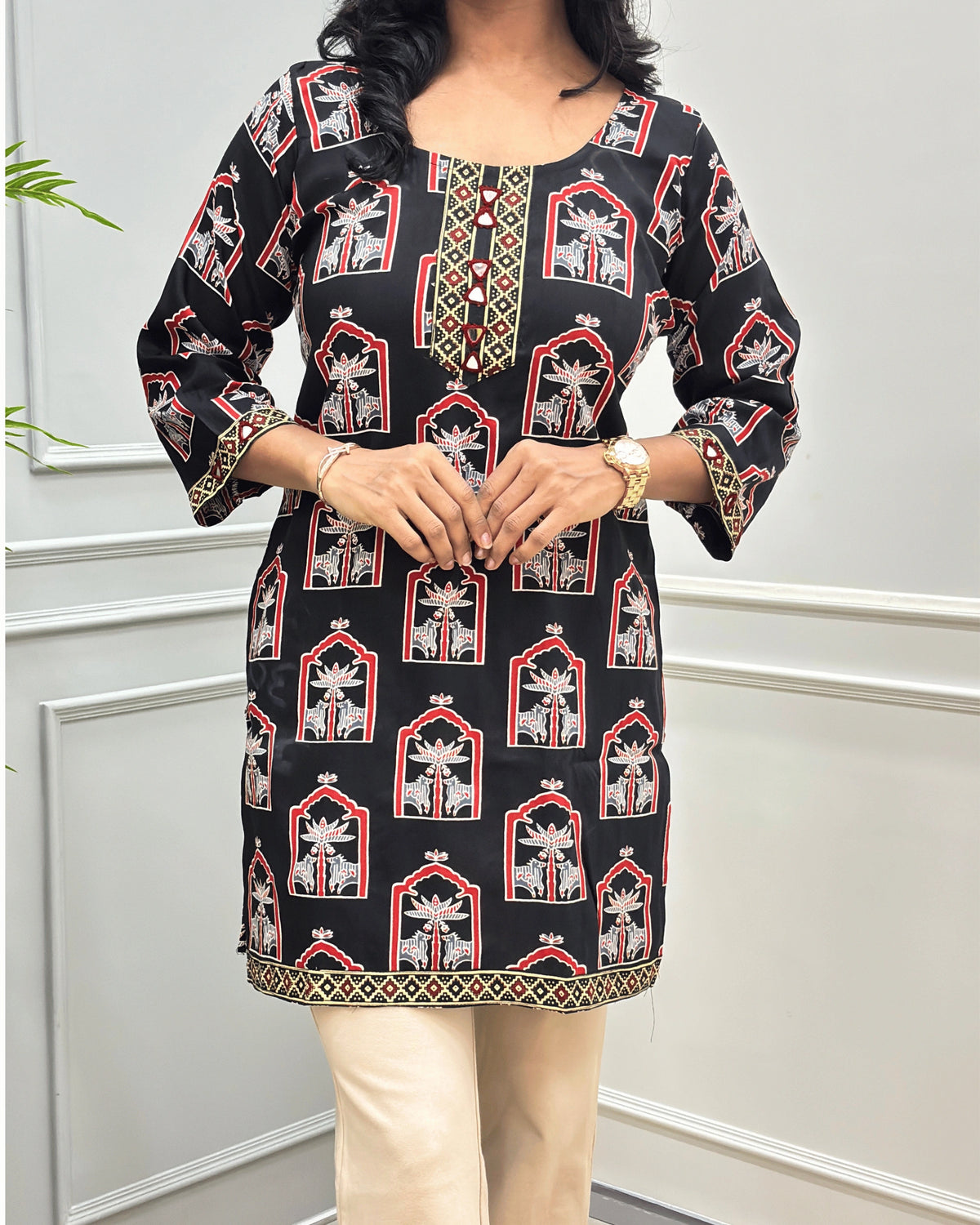 Close-up of Black women's Ajrakh plant based cotton short kurti with a traditional Indian arch pattern in black, white, and gold, hand-mirror work on front near Round neck , Ajrakh print on front, bottom sleeves cuffs , paired with beige flared pant , standing against a white paneled wall with a green plant on the left.