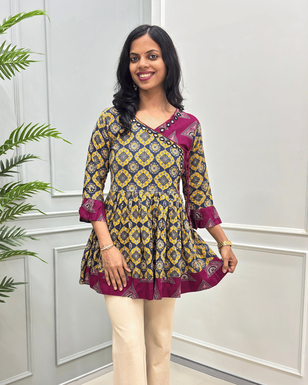 Front view of Mustard Yellow Ajrakh plant based rayon cotton Short Kurti featured with navy blue base & yellow geometric patterned with magenta borders, paired with beige pants, standing in front of a white paneled wall with a green plant on the left.