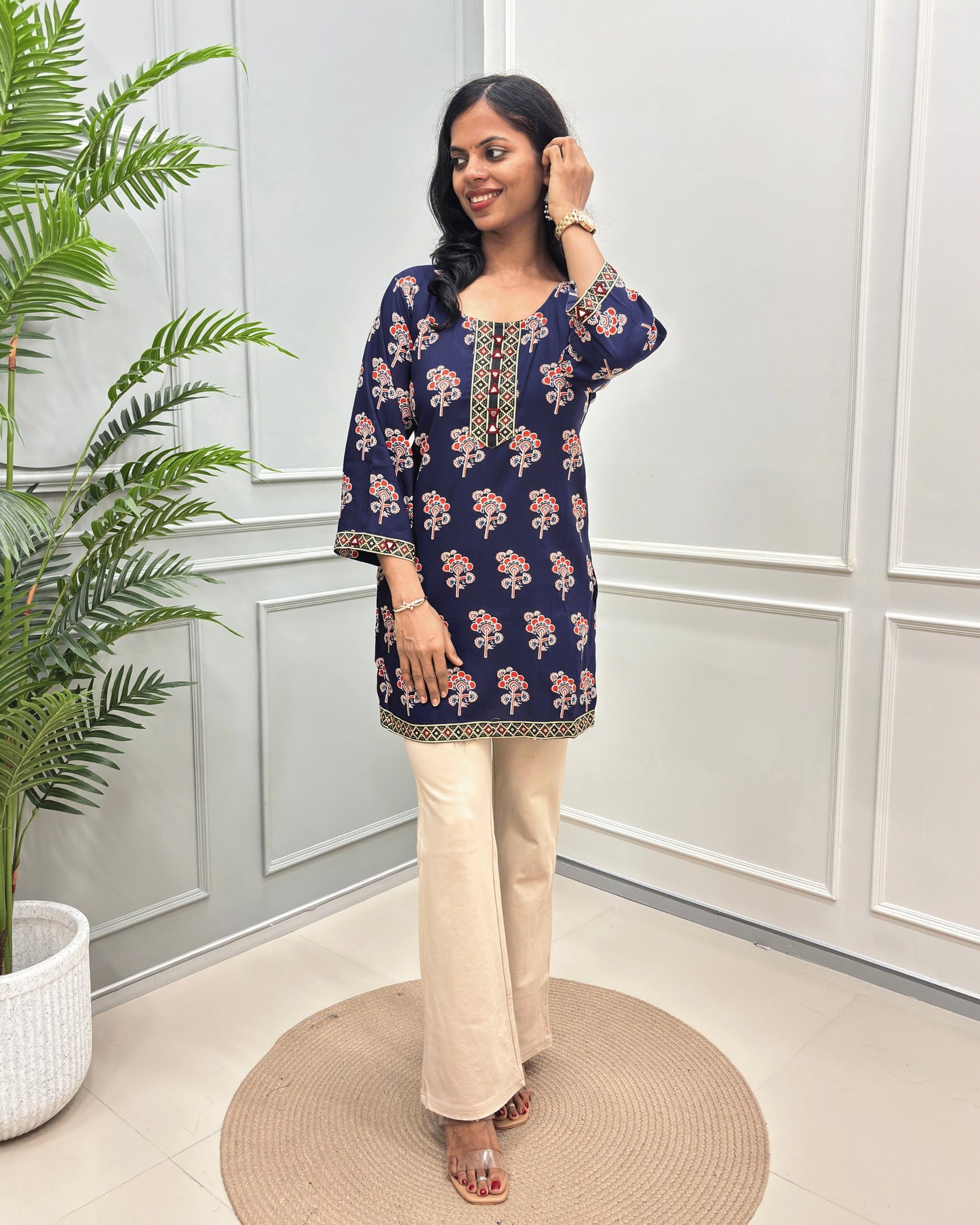 Navy-Blue Women's Ajrakh Plant based Rayon cotton Short kurti with a floral print , Ajrakh print on front, bottom & Sleeves beige wide-leg pants, standing on a round woven mat in front of a white paneled wall with a potted plant.