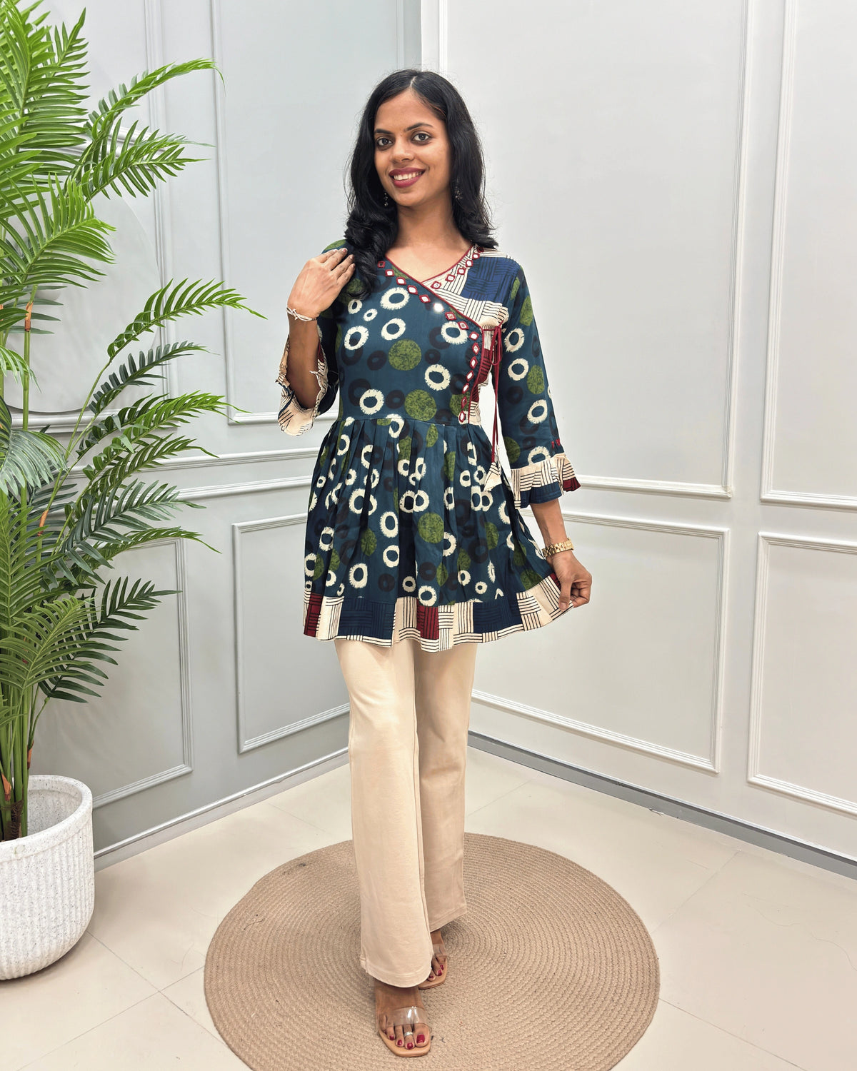 Green Women's Ajrakh Vegetable dyed plant based Cotton Short-Kurti with three-quarter frill sleeves, and waist to bottom,  paired with beige flared pant , standing on a round woven rug in front of a white paneled wall with a potted plant on the left.