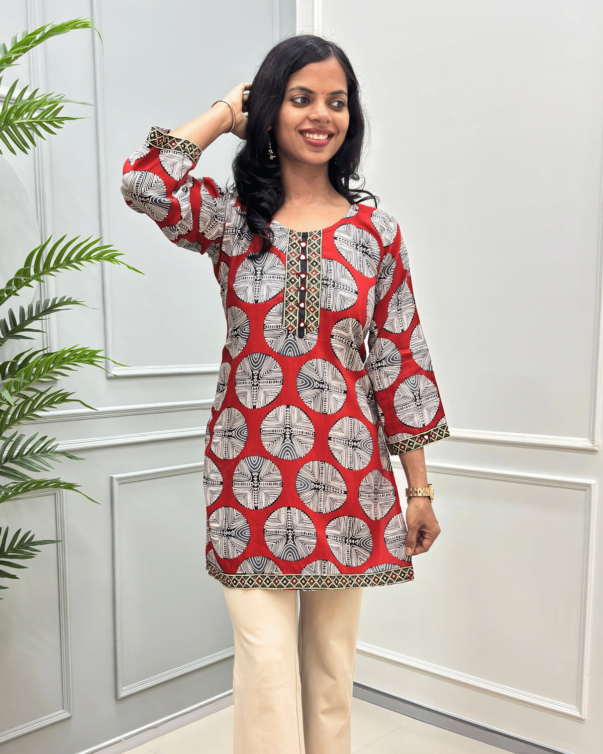 Semi-closeup of Red women's Vegetable dyed plant based Cotton Short kurti with a colorful geometric circular print and beige border trim & calf, paired with beige wide-leg trousers, standing on a round woven mat in front of a white paneled wall.