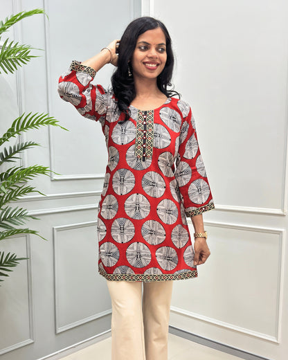 Semi-closeup of Red women's Vegetable dyed plant based Cotton Short kurti with a colorful geometric circular print and beige border trim & calf, paired with beige wide-leg trousers, standing on a round woven mat in front of a white paneled wall.
