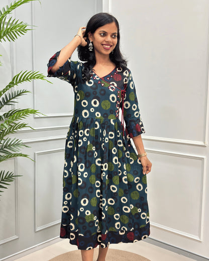 Green women's vegetable dyed plant based cotton long kurti front-view with a colorful white, green & black circle print,  and red floral accents, set against a white paneled wall with a green plant on the left.