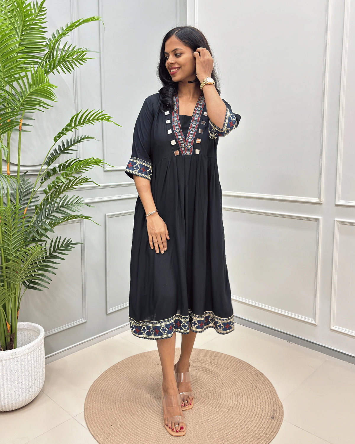 Black women's Vegetable dyed plant based Rayon cotton with colorful embroidered details on the neckline, sleeve cuffs, and hem, standing on a round woven mat in front of a white paneled wall with a potted plant.