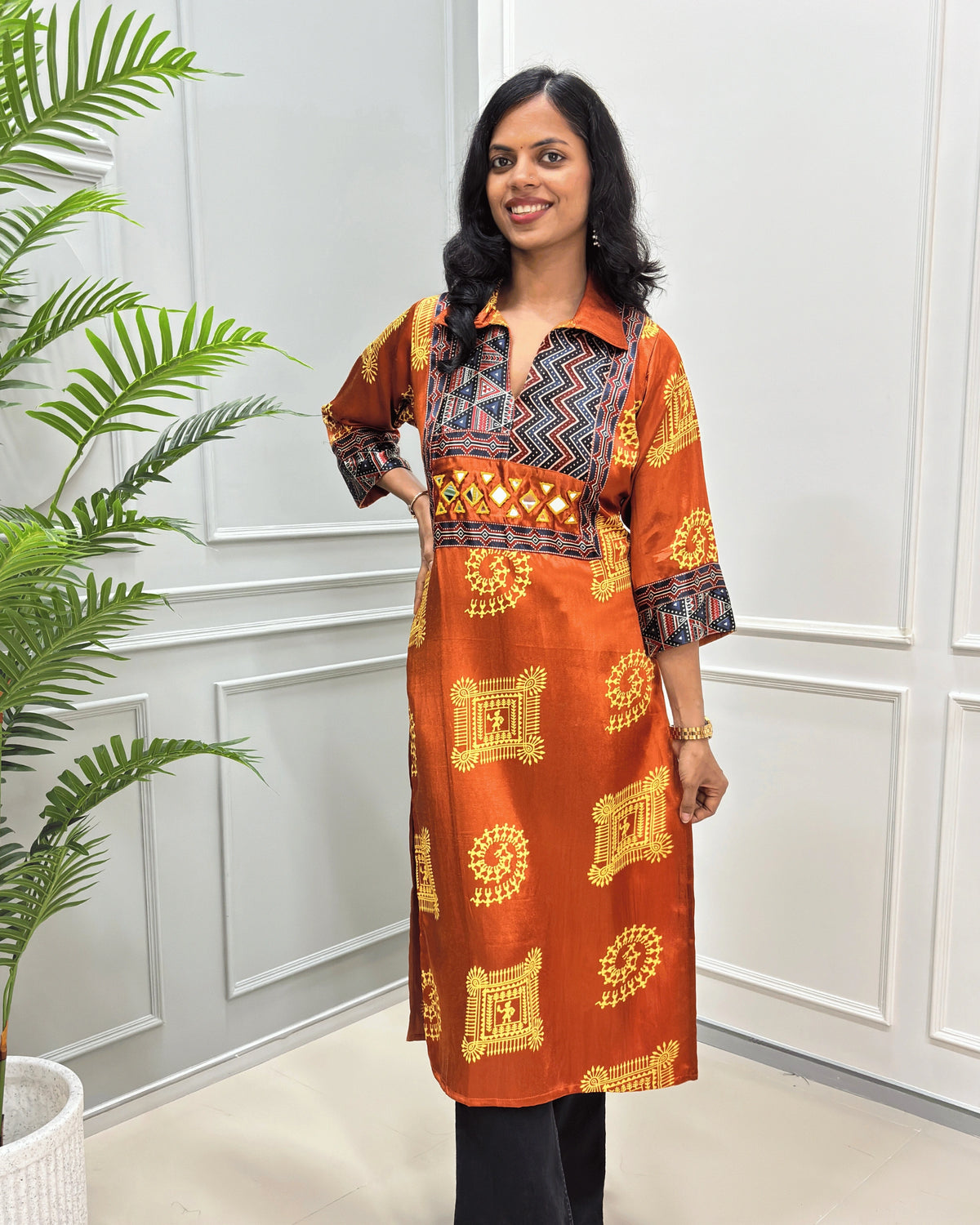 Front view of Orange Ajrakh pure Mashru Silk Long kurti with geometric print in red, white, and beige on Front & sleeves, standing in a bright room with a white paneled wall and a potted plant.