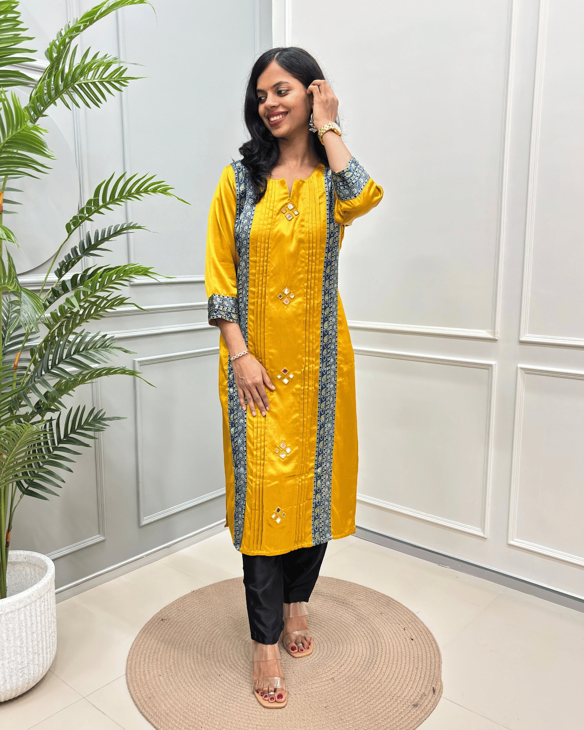 Yellow woman's Ajrakh Mashru Silk Long-Kurti with beige Ajrakh print panel on blue base on front and sleeves cuff and Hand-mirror work, paired with black pants, standing on a round woven rug in a modern indoor setting with a potted plant.