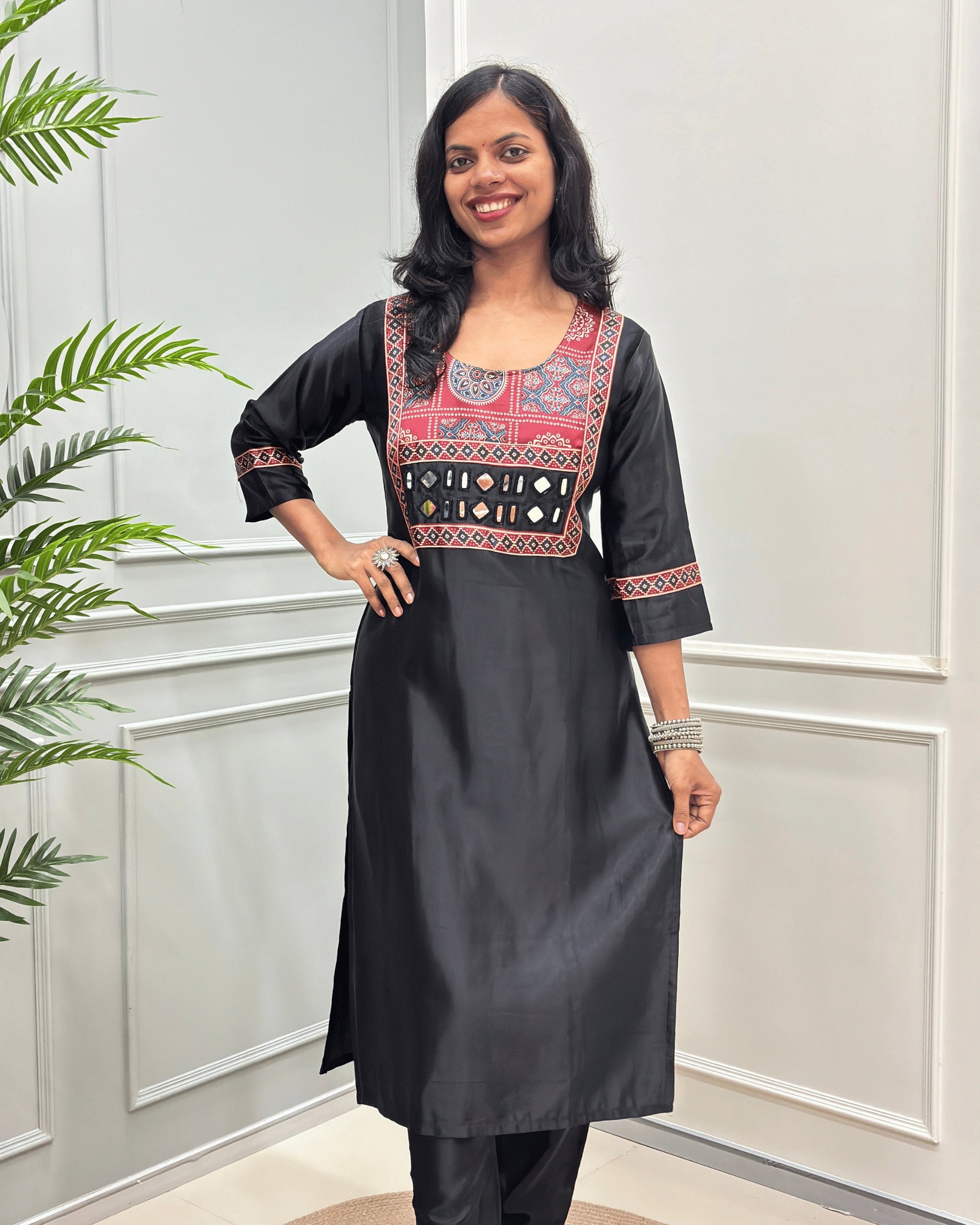 Front view of Black women's Ajrakh Mashru Silk Long-kurti with a blue, red, and white in Bandhani print , featuring Hand-mirror work with Yellow thread work embellishments , blue & red patterned cuffs, standing against a white paneled wall.