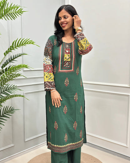Front View of Green women's 2 Peice Ajrakh Cotton Co-Ord Set with multi-color printed sleeves& Front, Along with Leaf pattern and a matching green Plazzo , standing in front of a white paneled wall with a green plant on the left.