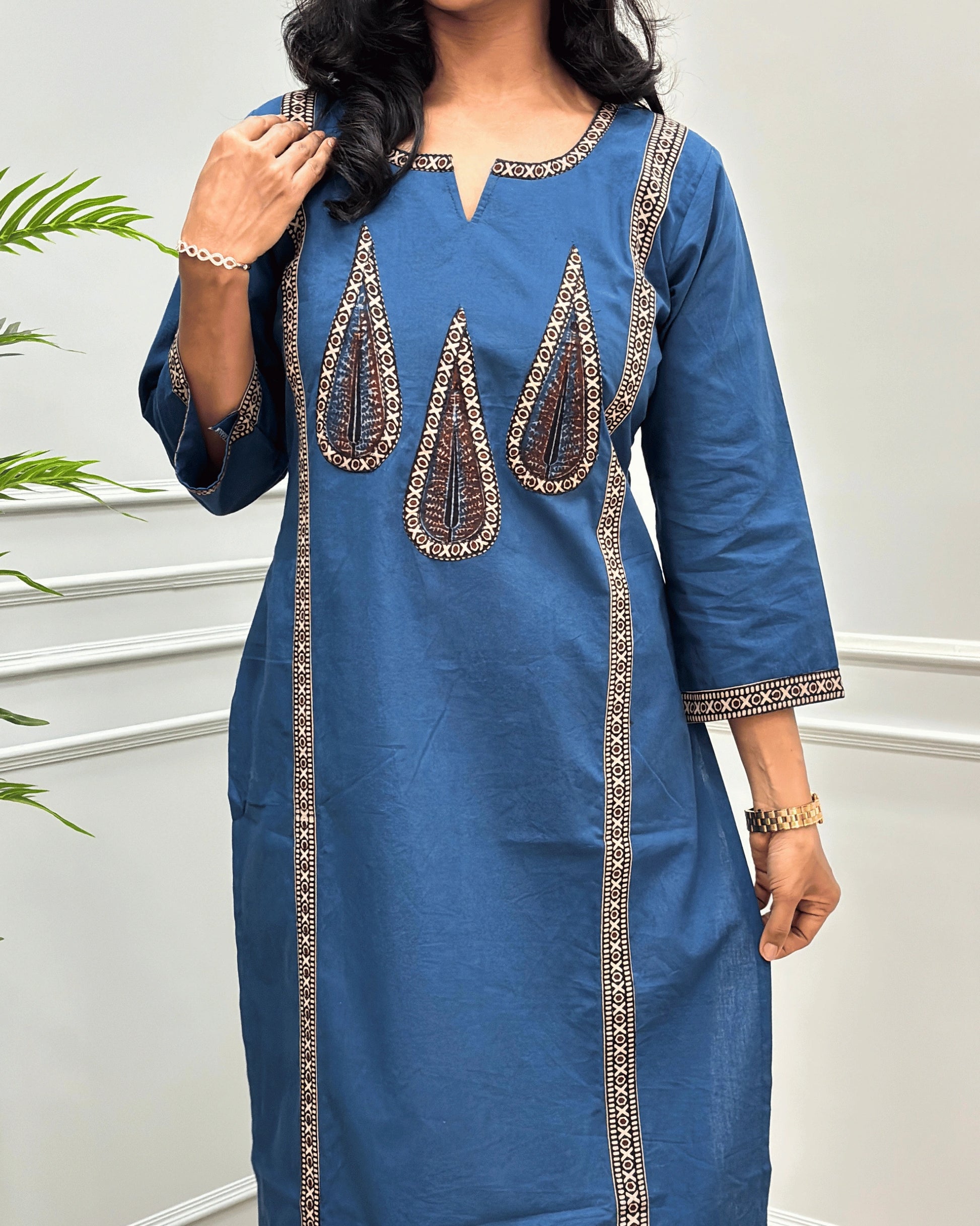 Close-up of Blue Women's Hand-block  Ajrakh Pure Cotton Long-kurti with three-quarter sleeve featured with intricate Beige , Black and brown embroidered details on the neckline, cuffs, and front  panel, set against a light gray wall with a plant in the background.