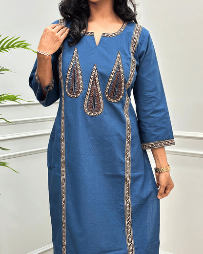 Close-up of Blue Women's Hand-block  Ajrakh Pure Cotton Long-kurti with three-quarter sleeve featured with intricate Beige , Black and brown embroidered details on the neckline, cuffs, and front  panel, set against a light gray wall with a plant in the background.
