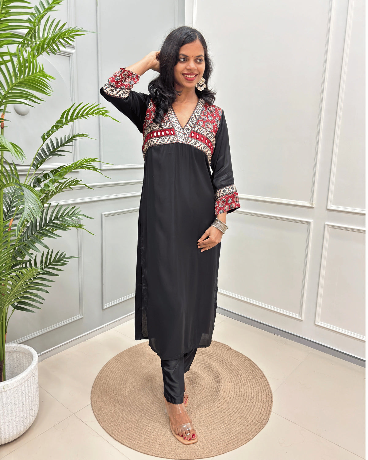Black Women's Ajrakh Modal Silk Long-Kurti with intricate Red base & White neck border , Ajrakh print on the yoke and cuffs, paired with black pants and clear heeled sandals, standing on a round woven mat in a decorated room.