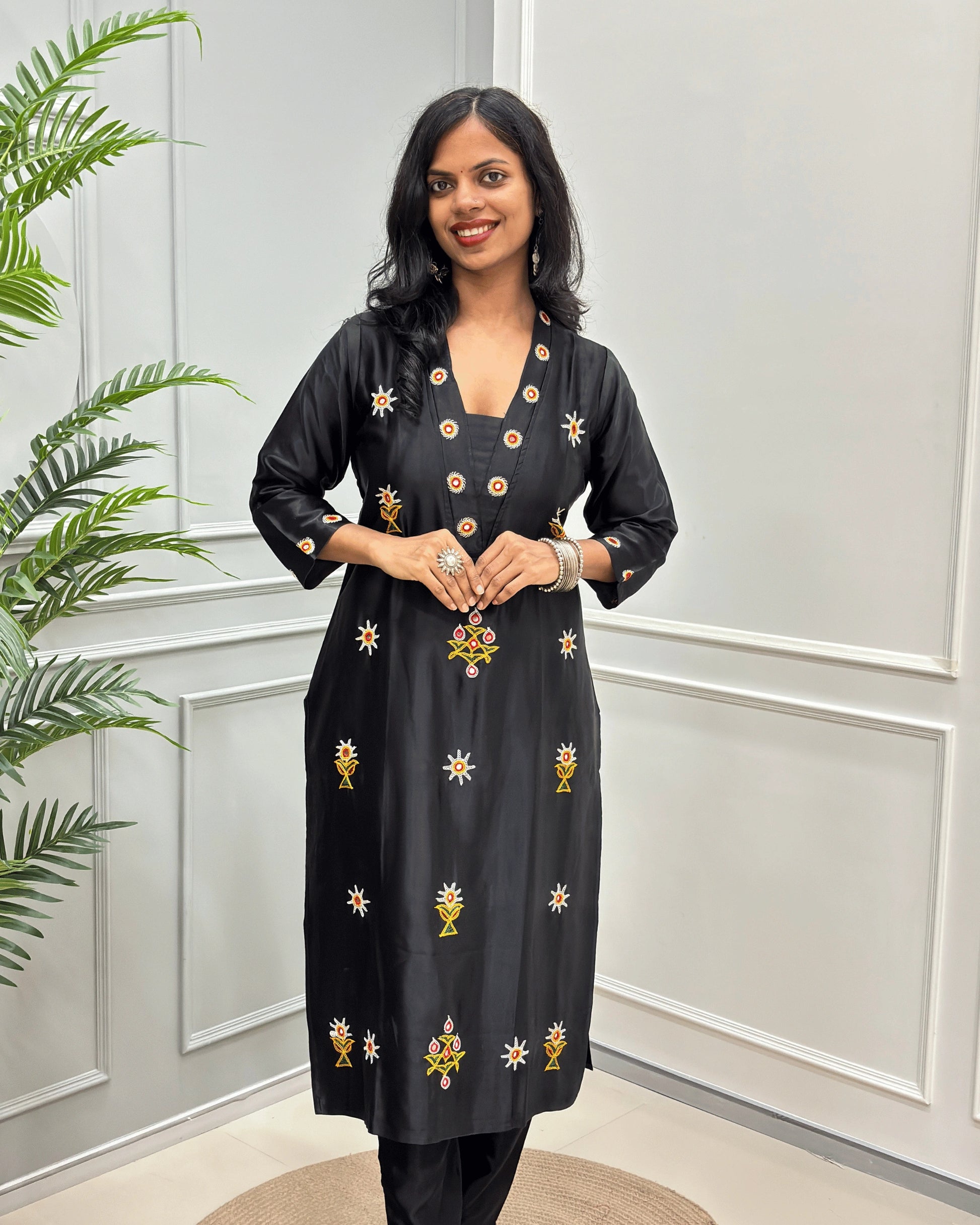 Front view of Black women's pure Mashru Silk with embroidered Ahir motifs, Hand-mirror work on front, V shaped neck, & Sleeves cuff,  standing in front of a white paneled wall with a green plant on the left.
