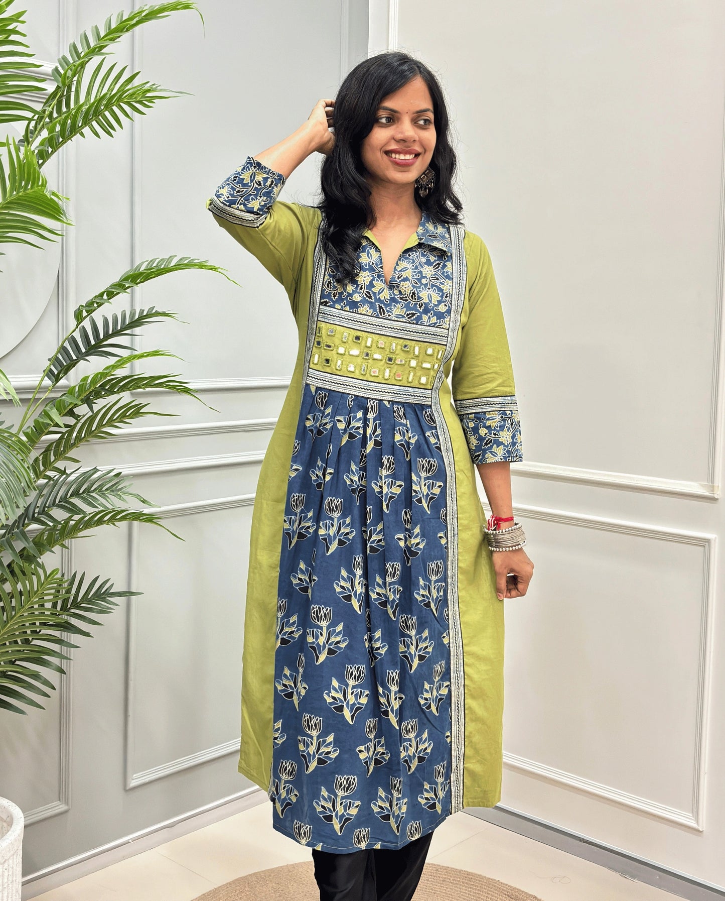Front view of Green women's Ajrakh plant based cotton Long-kurti featured with a Green outer kurta and a blue printed inner kurta with floral motifs on front & sleeves , set against a white paneled wall with a green plant on the left.
