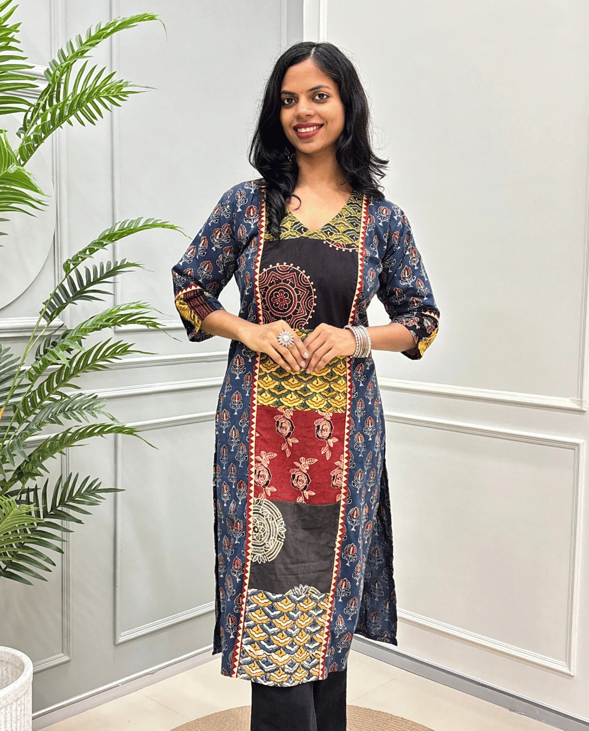 Front view of Blue women's Hand-block Ajrakh pure cotton long kurti with three quarter full printed sleeves with a navy blue base, featuring red, yellow, and black Ajrakh Geometric print panel & cuffs, standing in front of a light gray paneled wall with a green plant on the left.