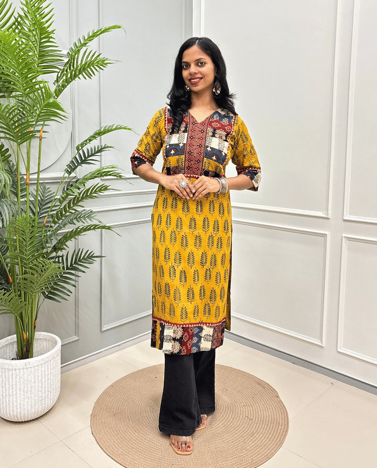 Yellow Women's Hand-block Ajrakh Pure cotton Long-Kurti with multicolor patchwork-style yoke, Cuffs and hem panels, paired with black Flared pants, standing indoors against a white paneled wall next to a potted plant.