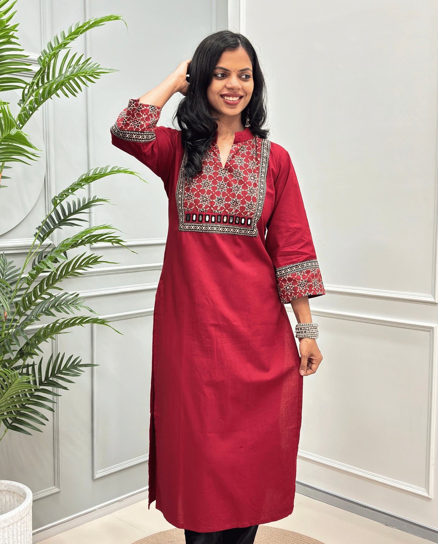 Front view of Red Women's Hand-block Ajrakh Pure Cotton Long kurti with intricate green geometric print on the chest and sleeve cuffs, hand-mirror work on front , paired with black pants , standing on a round woven mat in front of a white paneled wall with a potted plant.