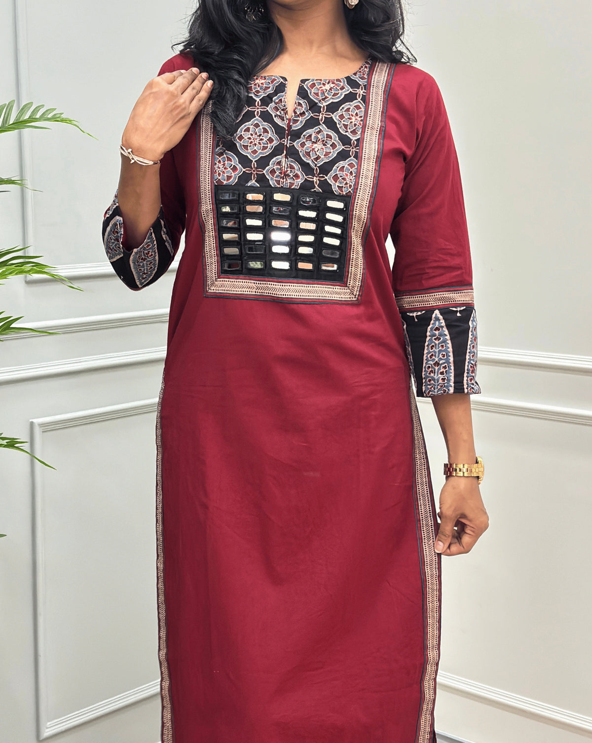 Close-up of Red Women's Hand-block Ajrakh Pure Cotton Long Kurti Featured with Three-quarter Sleeves & Round neck with V-shape cut with a Black base and abstract Red , Grey & Beige geometric embroidery on the chest and sleeve cuffs, Hand-mirror work on Front, Ajrakh print border on Sides & Front, with Side Pockets, paired with black Flared pants, standing in a decorated room with a potted plant.