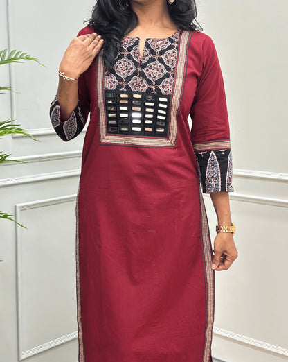 Close-up of Red Women's Hand-block Ajrakh Pure Cotton Long Kurti Featured with Three-quarter Sleeves & Round neck with V-shape cut with a Black base and abstract Red , Grey & Beige geometric embroidery on the chest and sleeve cuffs, Hand-mirror work on Front, Ajrakh print border on Sides & Front, with Side Pockets, paired with black Flared pants, standing in a decorated room with a potted plant.