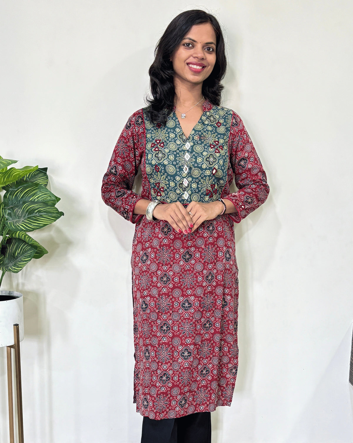 Front view of Red Women's Ajrakh Vegetable Dyed plant based cotton long kurti with green and red patterns, Hand-mirror work on front & Sleeves paired with black flared pants, standing in a room with a white wall and a potted plant.
