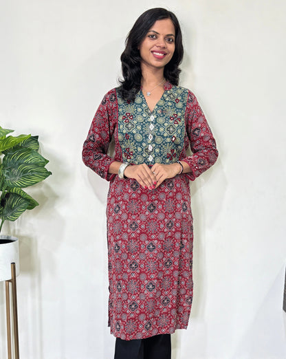 Front view of Red Women's Ajrakh Vegetable Dyed plant based cotton long kurti with green and red patterns, Hand-mirror work on front & Sleeves paired with black flared pants, standing in a room with a white wall and a potted plant.