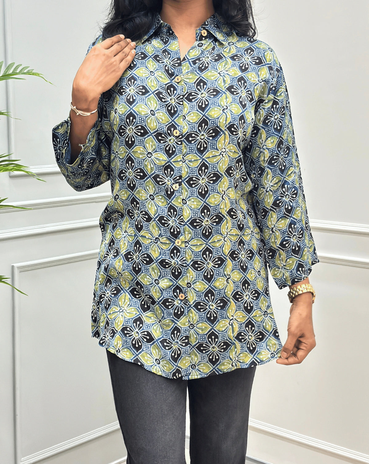 Close-up of Green Women's Ajrakh Pure Modal Silk Shirt with Full-Sleeves, with a Floral print on blue, yellow, and white geometric pattern, black flared pants, standing on a round woven rug in front of a white paneled wall with a potted plant on the left