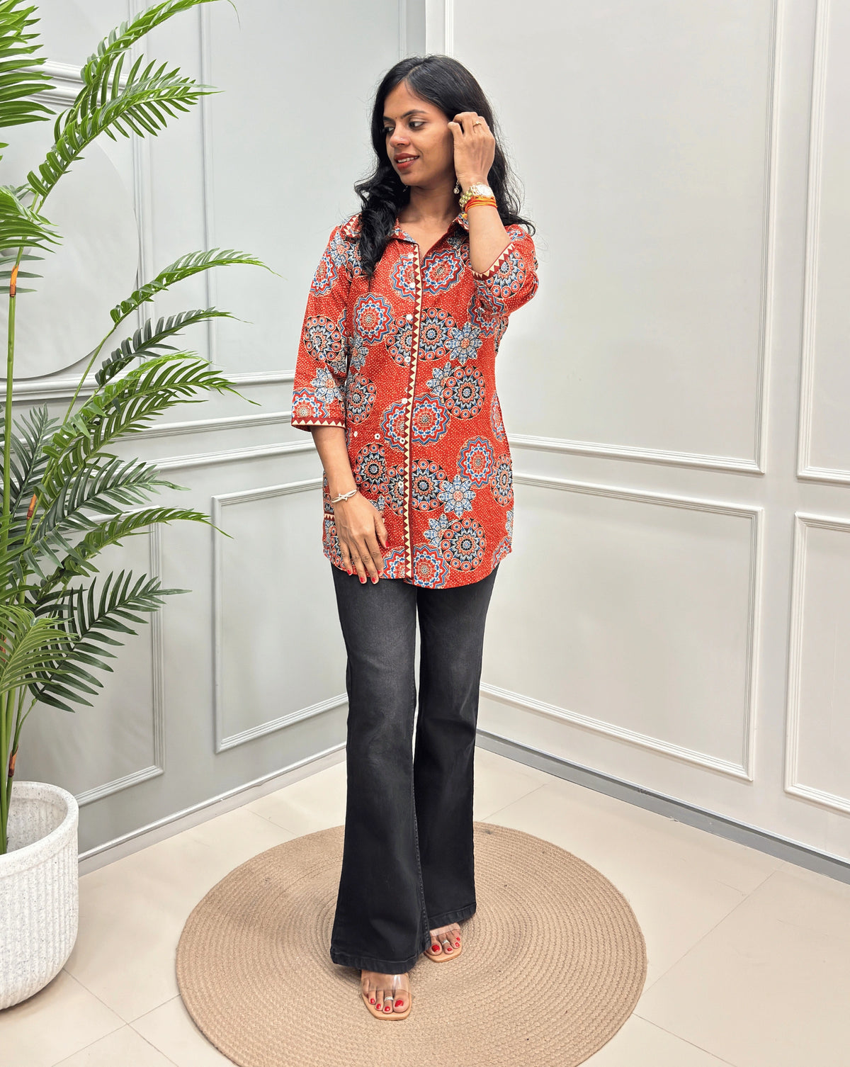 Orange women's Ajrakh pure cotton Shirt with a orange base and Multi color floral print,  paired with black flared pants. Standing on a round woven rug against a light paneled wall with a potted plant at left.