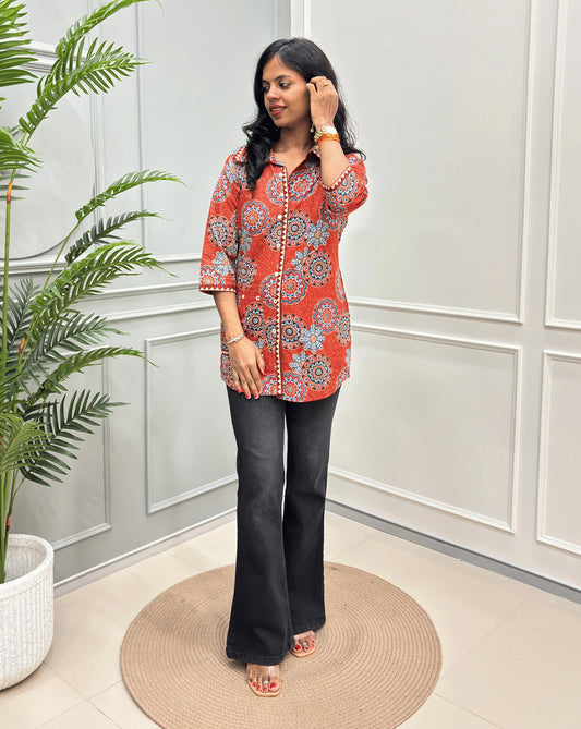 Orange women's Ajrakh pure cotton Shirt with a orange base and Multi color floral print,  paired with black flared pants. Standing on a round woven rug against a light paneled wall with a potted plant at left.