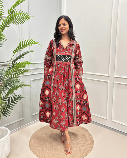 Woman's Red hand-block cotton long A-line dress with intricate black, beige, and blue floral patterns, and Ajrakh printed sleeves , standing on a round woven mat in front of a white paneled wall.