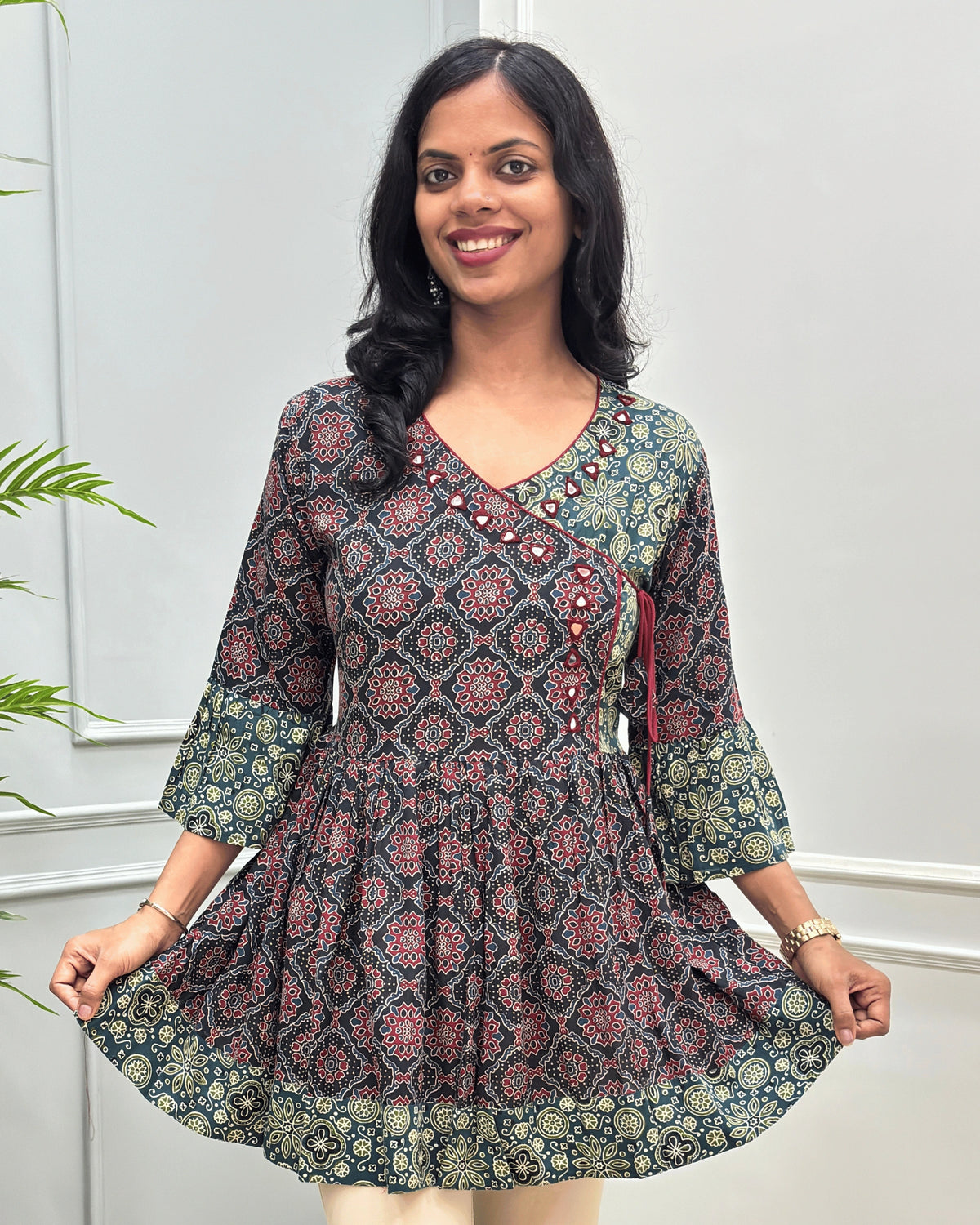 Close-up of Black women's Ajrakh Plant based cotton short-kurti with a Three-Quarter Sleeves (frill)  featuring Black & Green Geometric print ,  Angrakha Pattern with Hand-mirror work on V-shaped Neckline along with Lace, set against a light gray paneled wall.