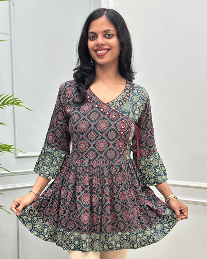 Close-up of Black women's Ajrakh Plant based cotton short-kurti with a Three-Quarter Sleeves (frill)  featuring Black & Green Geometric print ,  Angrakha Pattern with Hand-mirror work on V-shaped Neckline along with Lace, set against a light gray paneled wall.