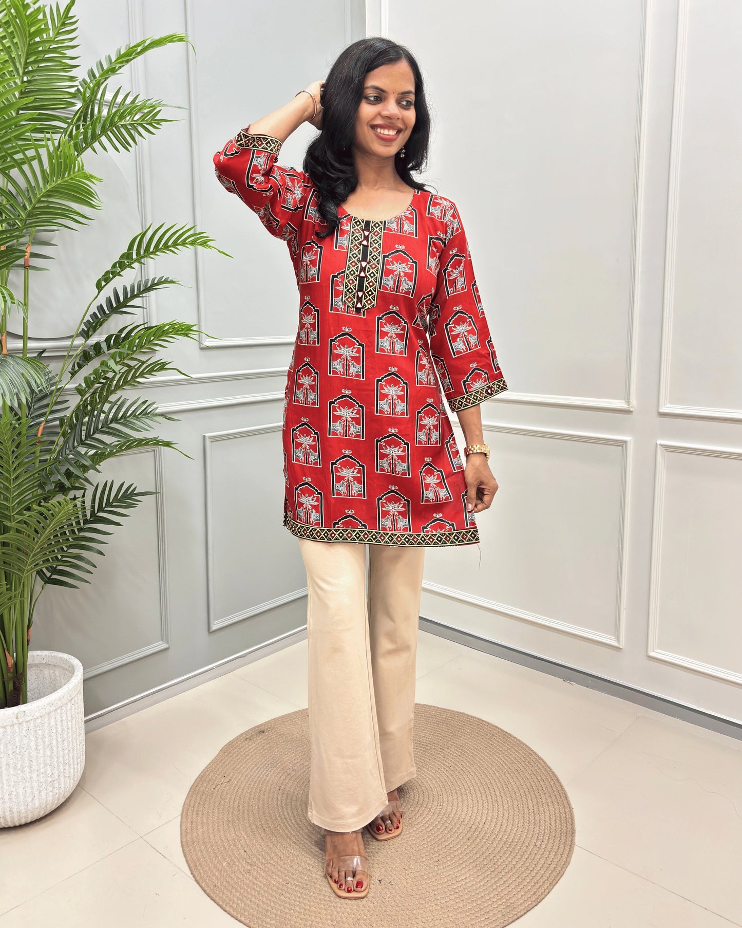 Red Women's Ajrakh vegetable Dyed Plant based Cotton Short Kurti with a colorful Pichhwai pattern, Paired with beige flared pants, standing on a round woven mat in front of a white paneled wall with a potted plant.