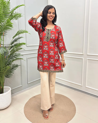 Red Women's Ajrakh vegetable Dyed Plant based Cotton Short Kurti with a colorful Pichhwai pattern, Paired with beige flared pants, standing on a round woven mat in front of a white paneled wall with a potted plant.