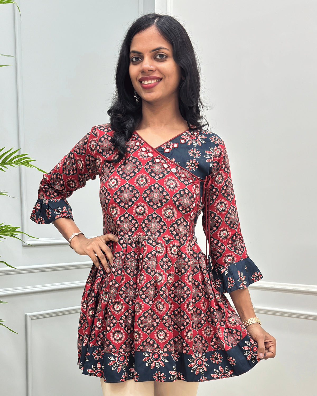 Close-up of Red plant based cotton  short kurti with three-quarter ruffled sleeves, beautiful Angrakha pattern with lace, hand-mirror work on V-neck standing against a light gray paneled wall
