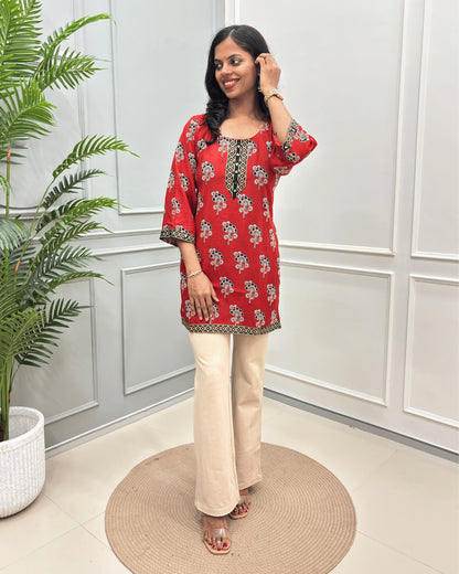 Red Women's Ajrakh Plant based Rayon cotton Short kurti with a floral print , Ajrakh print on front, bottom & Sleeves beige wide-leg pants, standing on a round woven mat in front of a white paneled wall with a potted plant.