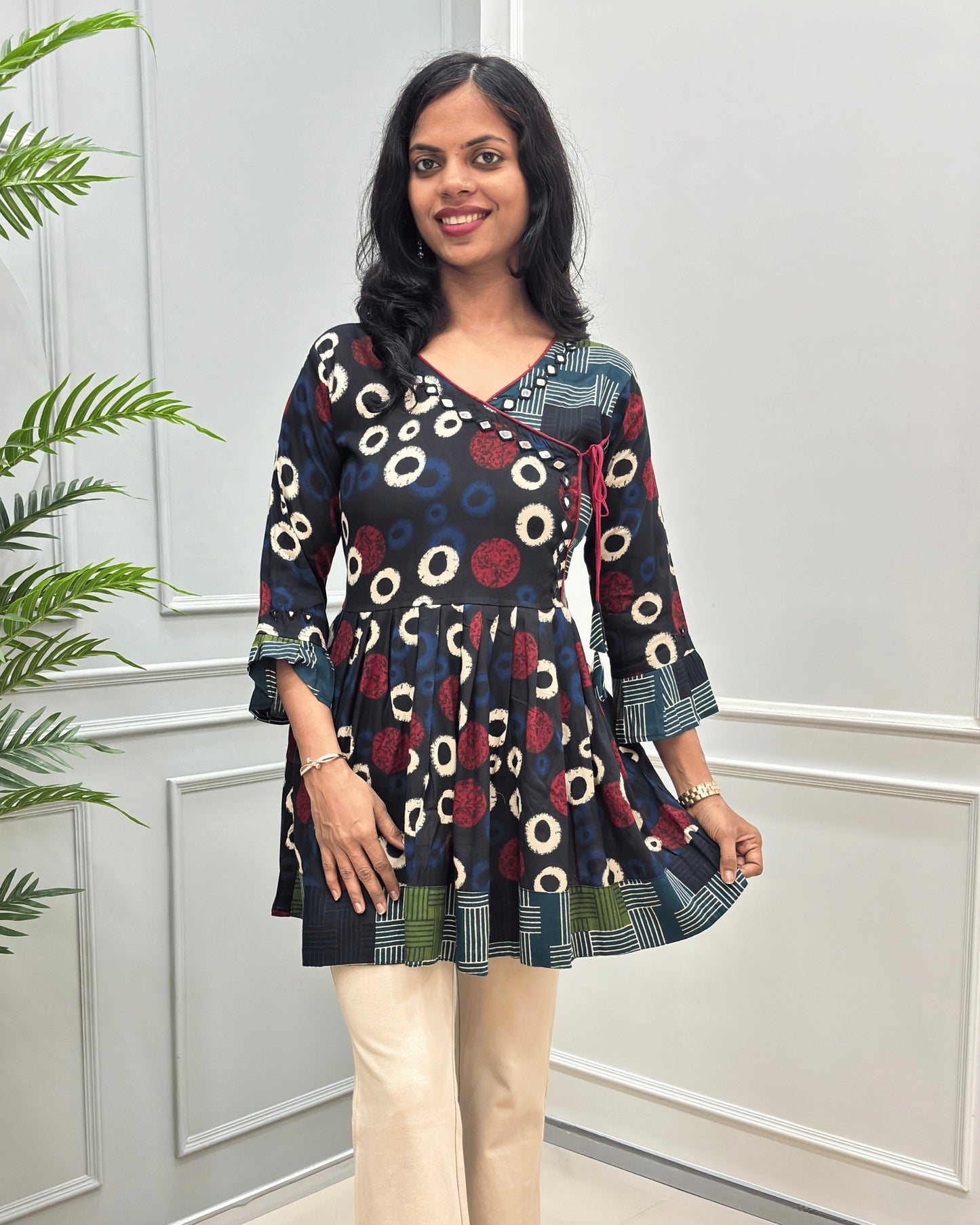Front view of Black women's Ajrakh Plant based cotton Short-Kurti with a multicolor abstract print of circles and geometric patterns, Strip print on top bottom & sleeves cuffs in paired with beige pants, standing in front of a white paneled wall.