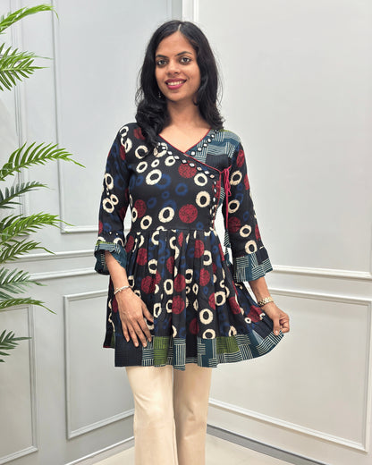 Front view of Black women's Ajrakh Plant based cotton Short-Kurti with a multicolor abstract print of circles and geometric patterns, Strip print on top bottom & sleeves cuffs in paired with beige pants, standing in front of a white paneled wall.