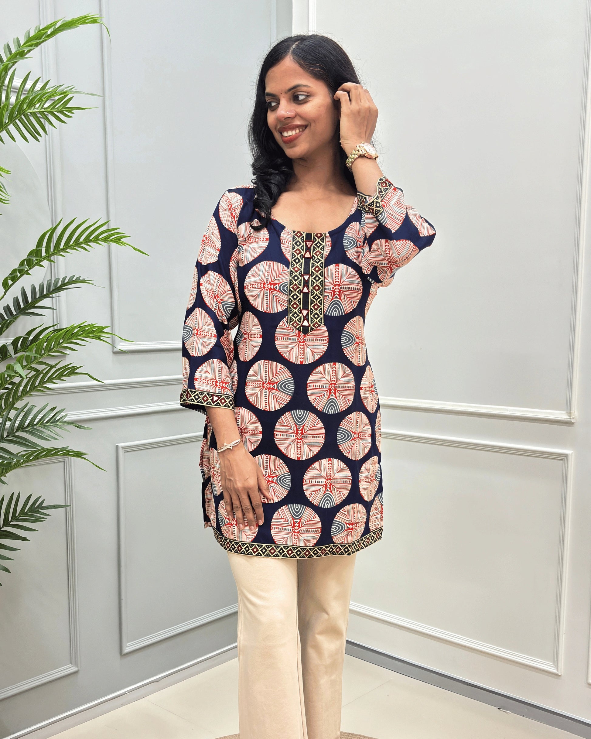 Semi-close-up of Navy-Blue Plant based cotton Short-kurti with multi-color geometric circular print and paired with beige pants, standing in a bright room with white paneled walls and a green plant on the left.