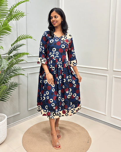 Blue Women's Vegetable dyed Plant based cotton long -kurti with a red, Black and cream abstract circle print, Angrakha pattern with lace standing on a round woven rug in front of a white paneled wall.