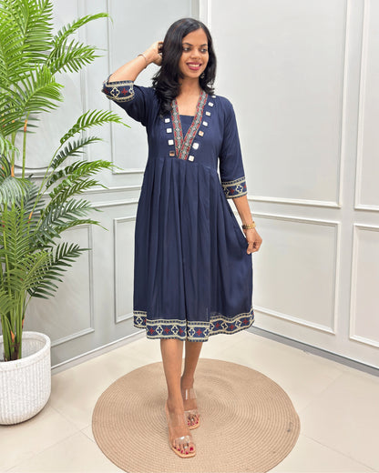 Navy-blue women's vegetable dyed plant based cotton A-line long-kurti with three quarter sleeves & calf length kurti with colorful geometric embroidery on the neckline, sleeve cuffs, and hem, standing on a round woven mat in front of a white paneled wall with a potted plant.