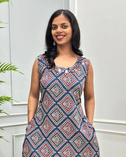 Close-up of Blue Women's cotton long sleeveless Kurti with a red, white, and beige geometric diamond pattern, Hand-mirror work on rounnd neck line border,  standing against a white paneled wall.