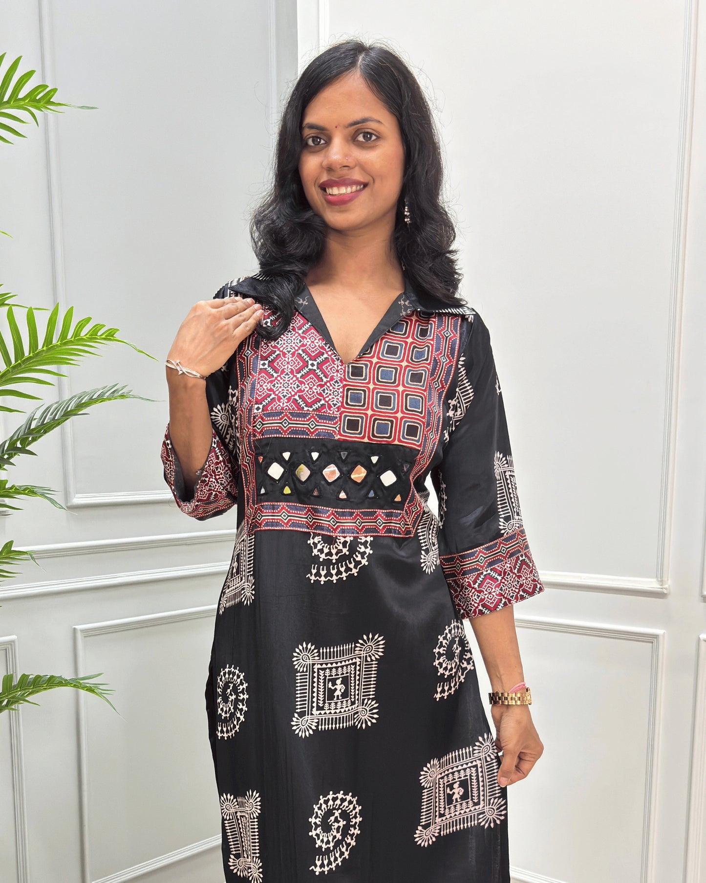 Close-up of Black Ajrakh Mashru silk Long kurti with multicolor geometric , warli print on front & sleeves along eith hand -mirror work on front , Collar V shaped neck patterns, standing in front of a white paneled wall with a green plant on the left.
