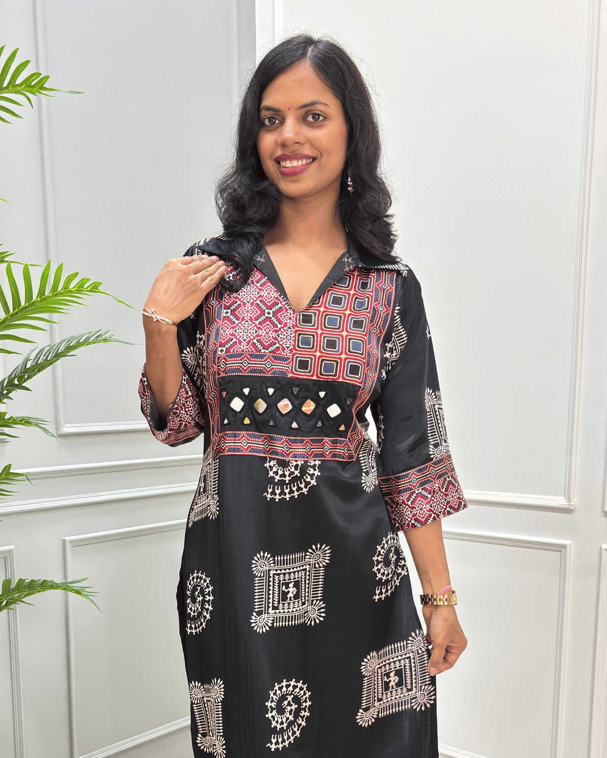 Close-up of Black Ajrakh Mashru silk Long kurti with multicolor geometric , warli print on front & sleeves along eith hand -mirror work on front , Collar V shaped neck patterns, standing in front of a white paneled wall with a green plant on the left.