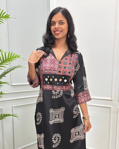 Close-up of Black Ajrakh Mashru silk Long kurti with multicolor geometric , warli print on front & sleeves along eith hand -mirror work on front , Collar V shaped neck patterns, standing in front of a white paneled wall with a green plant on the left.