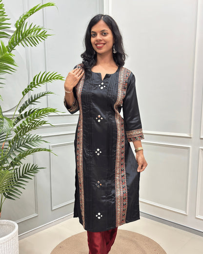 Front view of Black women's Ajrakh Mashru Silk Long Kurti with ornate multicolor embroidered Vertical  patterned on front & sleeves cuffs , paired with maroon pants, standing in a bright room with a white paneled wall and a green plant.