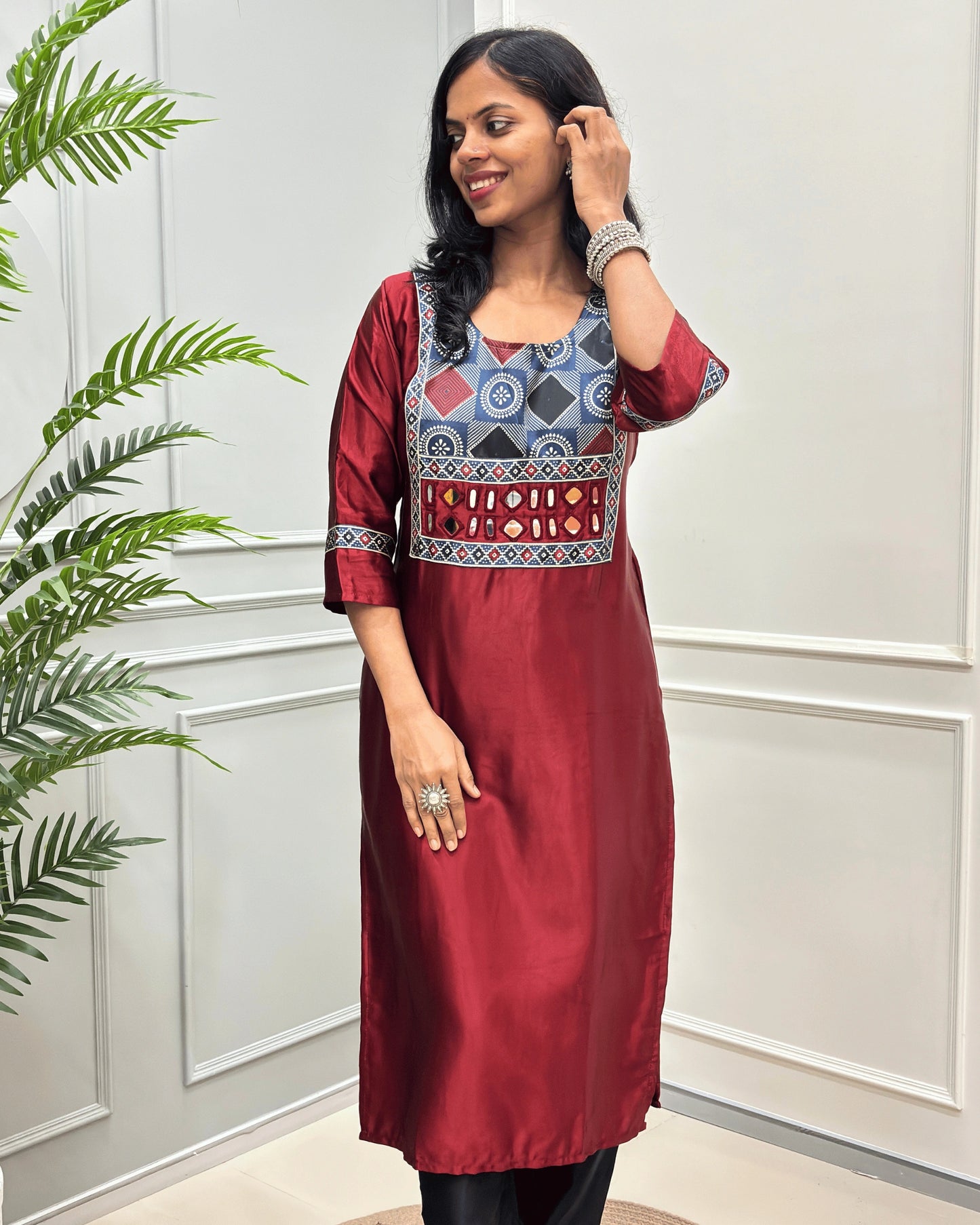 Front view of Red Women's  Ajrakh Mashru Silk Long-kurti with a blue, red, and white in Bandhani print , featuring Hand-mirror work with Yellow thread work embellishments , blue & red patterned cuffs, standing against a white paneled wall.