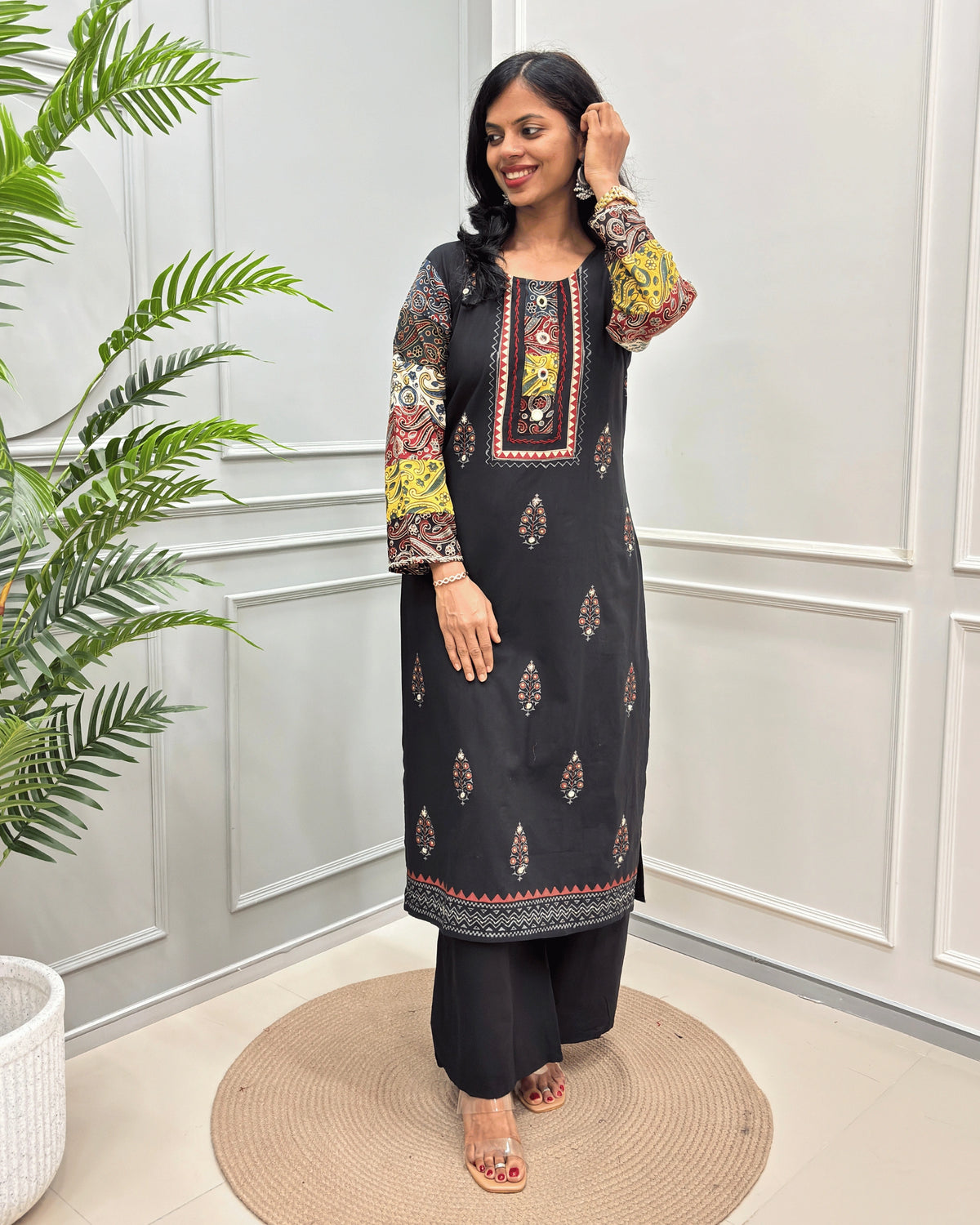 Black Women's Ajrakh Plant Based Rayon Cotton Co-Ord Set with colorful printed sleeves and embroidered details, standing on a round woven mat in front of a white paneled wall with a potted plant.