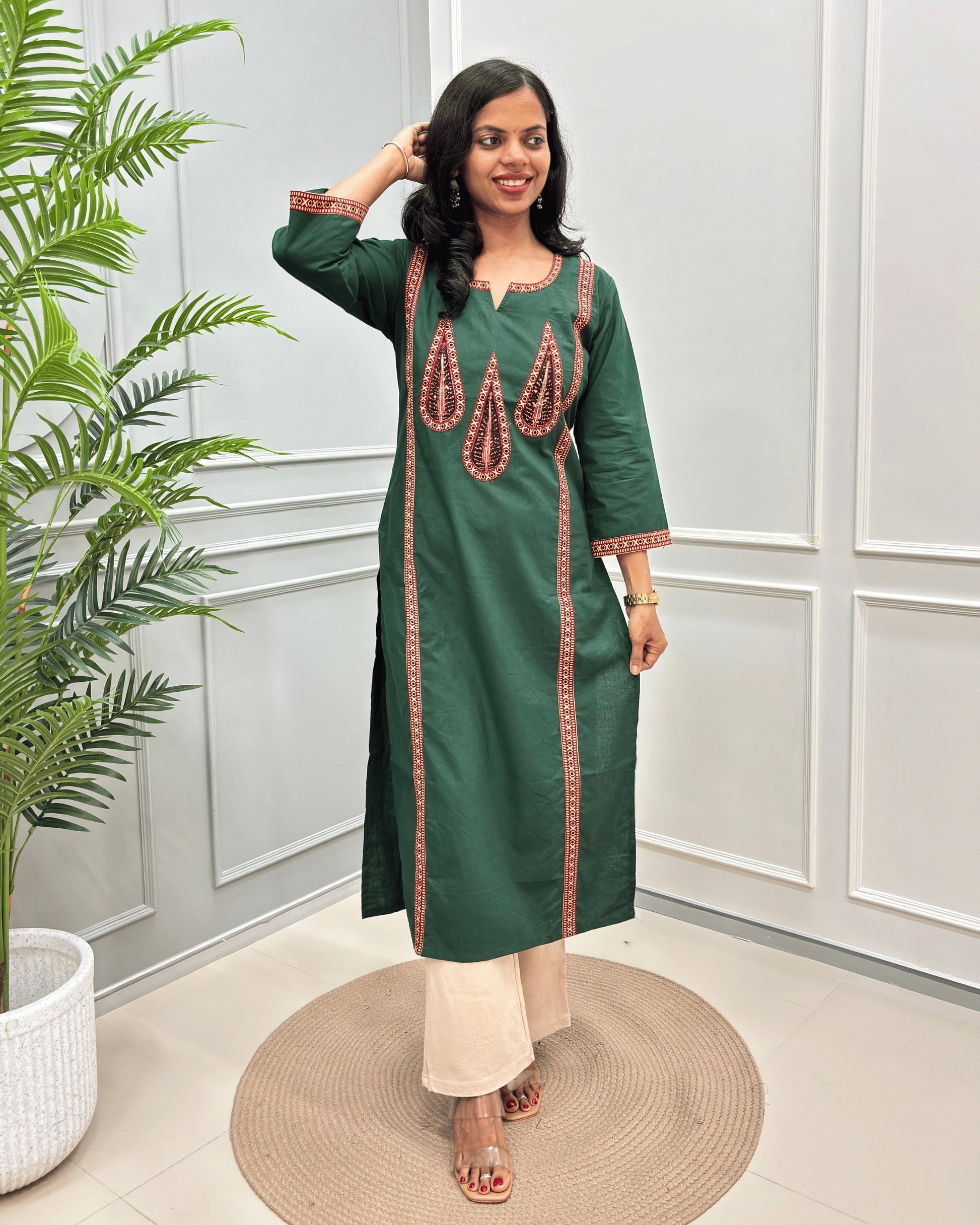 Front-view of Green Women's Hand-block Ajrakh Pure Cotton Long Kurti with Red and Black embroidered details on the V-neckline and borders on front & Sleeves , standing in a decorated room with a potted plant.