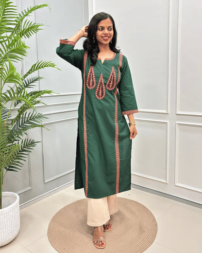 Front-view of Green Women's Hand-block Ajrakh Pure Cotton Long Kurti with Red and Black embroidered details on the V-neckline and borders on front & Sleeves , standing in a decorated room with a potted plant.