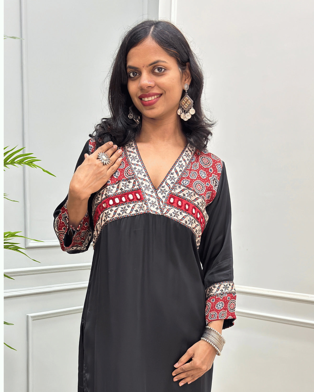 Close-up of Black Ajrakh Model silk fabric with three-quarter sleeves with intricate Red and Black patterned embroidery on the V-neckline, chest panel, and cuffs, standing in a light-colored room. 