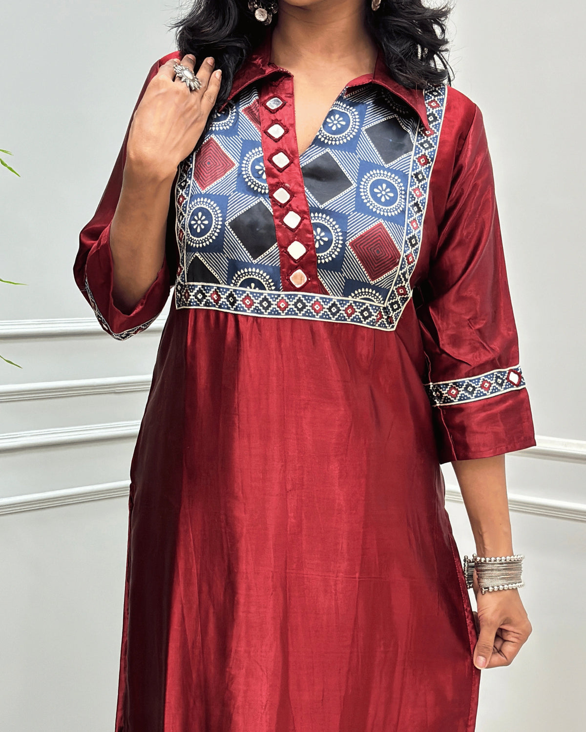 Close-up of Red Ajrakh Mashru Silk Long-kurti with a heavily embroidered Geometric yoke and patterned sleeves, Hand-mirror Work on Front & Sleeves, Collar V- neck,  standing against a light gray paneled wall.